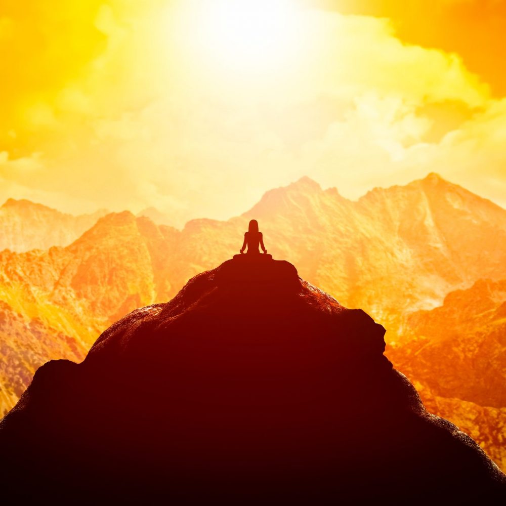 Woman meditating in sitting yoga position on the top of a mountains above clouds at sunset.